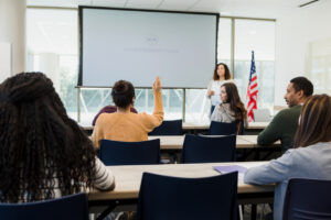 A rear view of a classroom full of diverse students taking an American citizenship class. One of the students, a woman, raises her hand to ask a question.