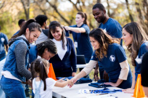 While other people register, a mid adult volunteer reaches across the table to shake hands with a young girl. Other volunteers smile and watch.